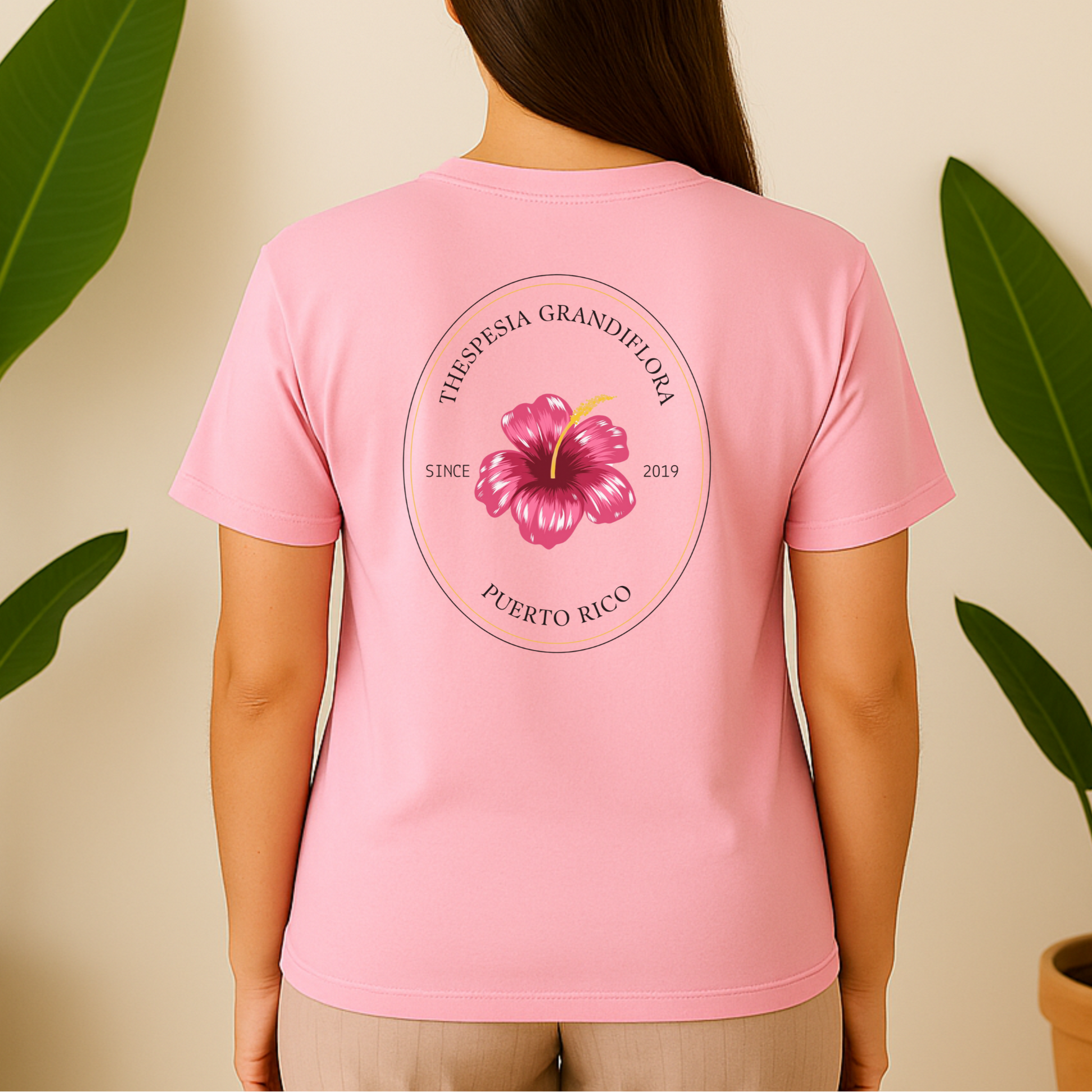 Person wearing a pink t-shirt with a floral design and text, standing against a beige wall with plants. Flor de Maga. Puerto Rico.
